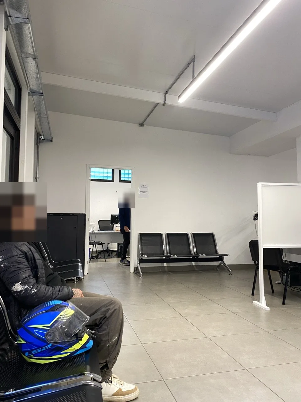 Зал ожидания отдела прав: ряды сидений, дверь с табличкой SALA TEÓRICOS, справа кабинки с компьютерами; лица двух присутствующих закрыты мозаикой