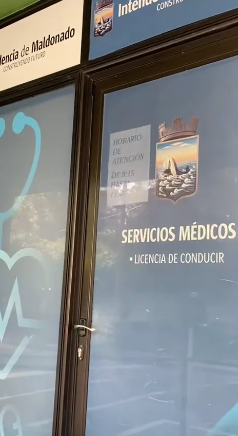 Стеклянная дверь муниципального медпункта с надписью SERVICIOS MÉDICOS • LICENCIA DE CONDUCIR и листком с часами приёма